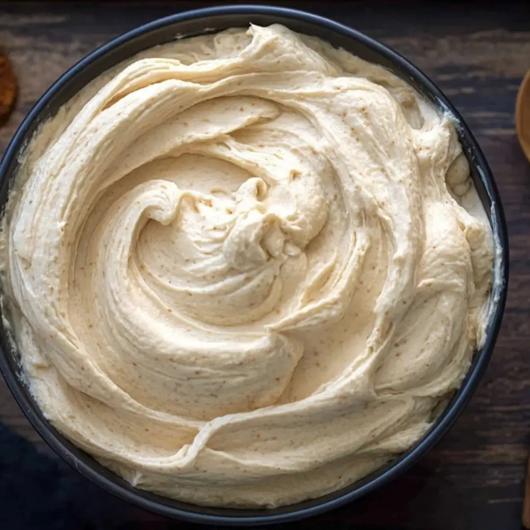 A top-down view of a black bowl filled with creamy, swirled Molasses Cream Cheese Frosting showing a light amber tint and spice flecks.
