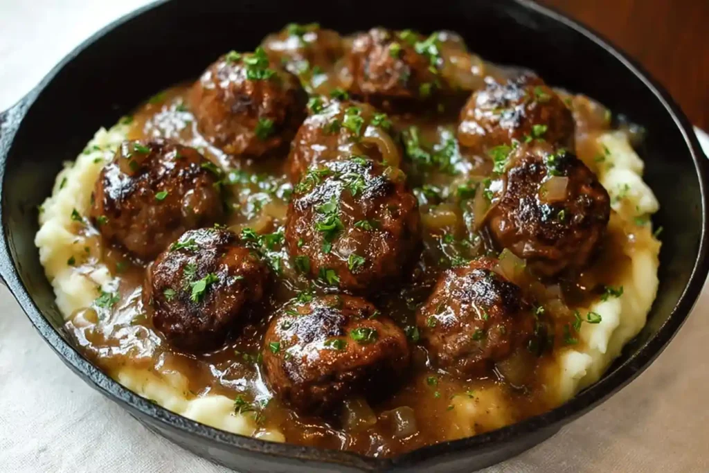 A close-up of Salisbury Steak Meatballs in a rich onion gravy, served over mashed potatoes in a black cast-iron skillet.