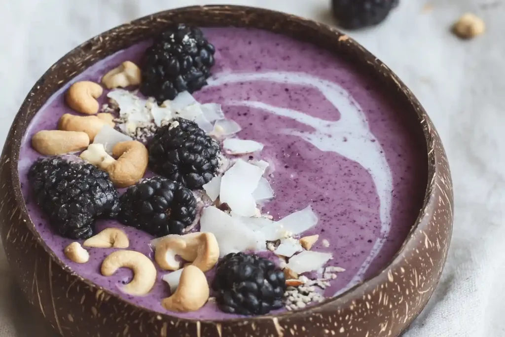 Top-down view of a purple blackberry smoothie bowl in a coconut shell topped with cashews and coconut.