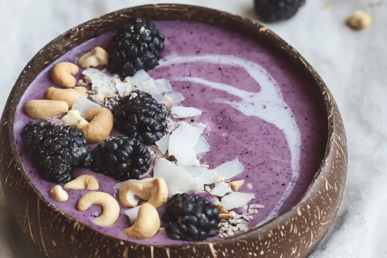 Top-down view of a purple blackberry smoothie bowl in a coconut shell topped with cashews and coconut.