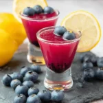 A vibrant blueberry lemon shot garnished with fresh berries and a lemon wheel on a dark slate board.