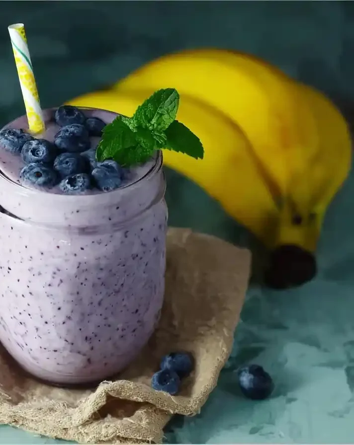 A glass jar filled with a thick blueberry smoothie, topped with fresh berries and mint, next to bananas and a lemon half.