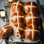 Freshly baked hot cross buns with icing crosses and raisins on a cooling rack next to butter.