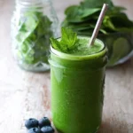 A vibrant spinach blueberry smoothie in a mason jar, garnished with fresh mint, surrounded by spinach leaves.