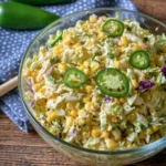 A glass bowl of creamy jalapeno corn coleslaw garnished with fresh pepper slices on a rustic wooden table.