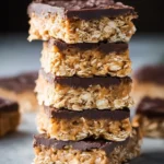 A tall stack of chewy peanut butter chocolate oat bars with a thick layer of dark fudge on top.