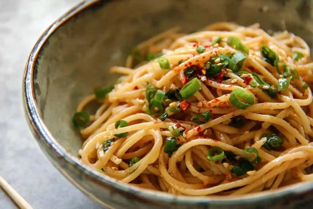 Close-up of a bowl of Sesame Noodles garnished with chopped green onions and red chili flakes
