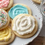 Close-up view of soft frosted sugar cookies on a white plate topped with pink, blue, yellow, and white buttercream.