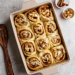 A baking dish filled with freshly baked apple cinnamon rolls topped with icing, next to cinnamon sticks.