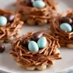 Close-up of four bird's nest cookies with scalloped edges, chocolate noodles, and candy eggs on a plate.