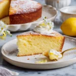 A slice of moist lemon cake on a plate, topped with whipped cream, powdered sugar, and small white flowers.
